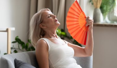 Stressed annoyed old senior woman using waving fan suffer from overheating, summer heat health hormone problem, no air conditioner at home sit on sofa feel exhaustion dehydration heatstroke concept
