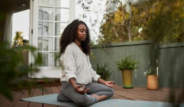 Young African American woman sitting on exercise mat outside on her patio and meditating in the lotus pose during a yoga session