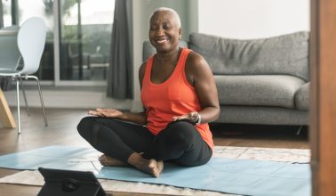 A beautiful senior woman takes an online yoga class. She is interacting with the teacher on her tablet and about to commence her class. She is wearing casual active wear and is taking the class in her lounge room. She is sitting on a blue yoga mat.