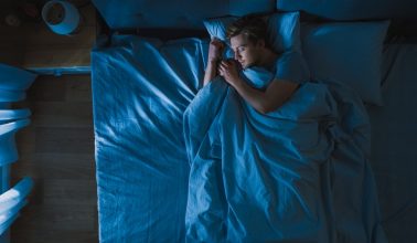 Top View of Handsome Young Man Sleeping Cozily on a Bed in His Bedroom at Night. Blue Nightly Colors with Cold Weak Lamppost Light Shining Through the Window.